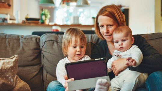 Mom sitting on the couch with toddler and baby looking at a tablet
