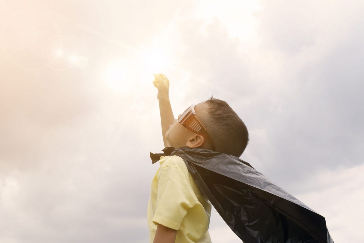 little boy with fist in the air looking at sky with a super hero cape and glasses on