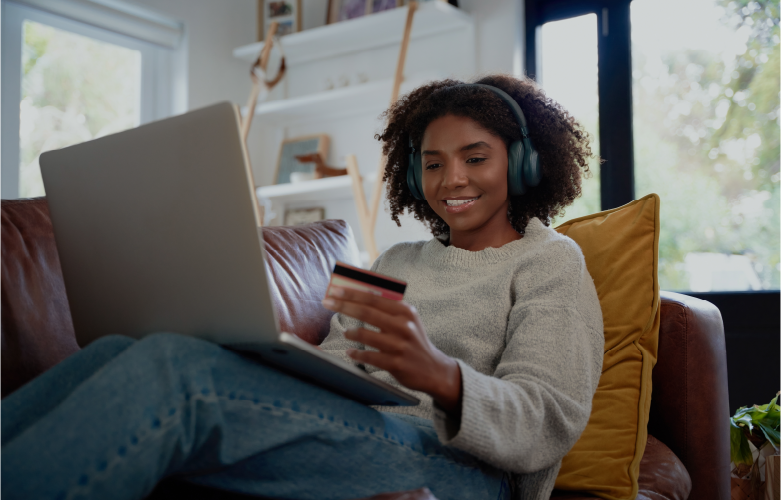young adult female sitting on couch with headphones on making online purchase with credit card