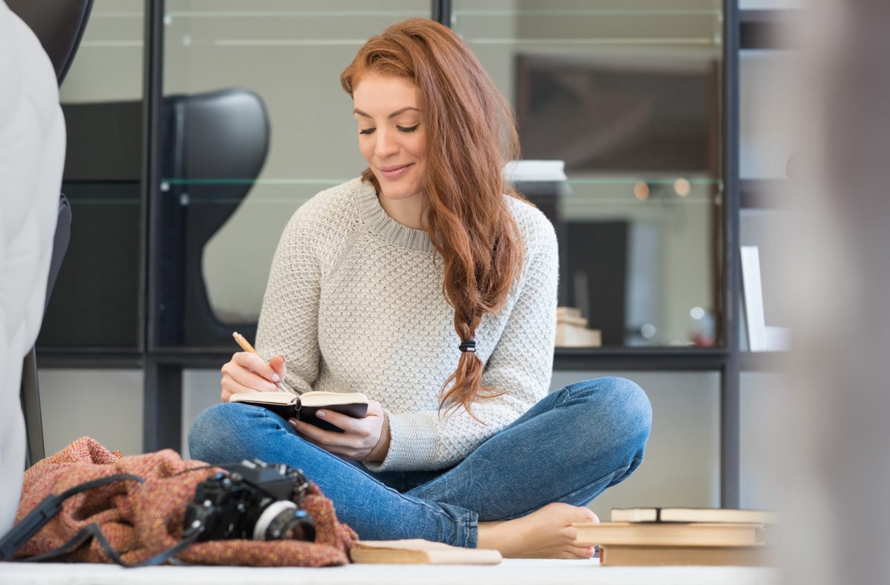 Girl with long red hair sitting on the floor with crossed legs writing in a book.